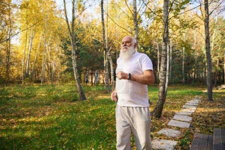 Téléchargez les photos : Vieil homme à barbe grise sur un jogging matinal près d'une maison de campagne, il est dans des vêtements légers confortables - en image libre de droit
