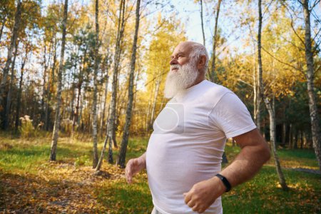 Téléchargez les photos : Vieil homme joyeux sur un jogging matinal dans une clairière forestière, il est dans des vêtements légers confortables - en image libre de droit