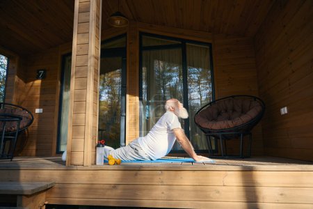 Viejo barbudo gris está haciendo yoga al aire libre, se encuentra en una terraza de madera