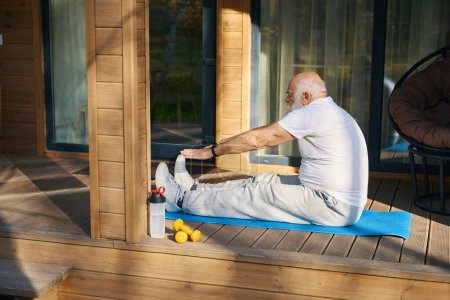 Téléchargez les photos : Le vieil homme à barbe grise fait des exercices matinaux dans l'air frais, il s'assoit sur une terrasse en bois - en image libre de droit