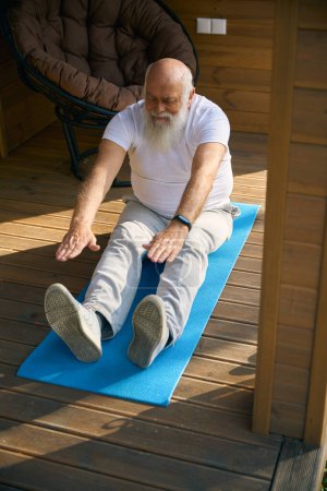 Téléchargez les photos : Homme âgé est engagé dans des exercices physiques sur la véranda d'une maison privée, il utilise un karimat - en image libre de droit
