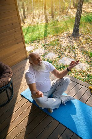 Photo for Elderly man sits in a yoga pose on the veranda of a private house, he uses a karimat - Royalty Free Image