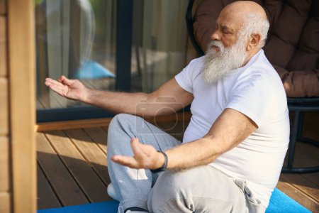 Téléchargez les photos : Vieil homme assis dans une pose de yoga sur une véranda en bois, il utilise un karimat - en image libre de droit