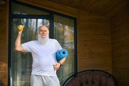 Cheerful pensioner with dumbbells and a gym mat on the veranda, he is working out in the fresh air