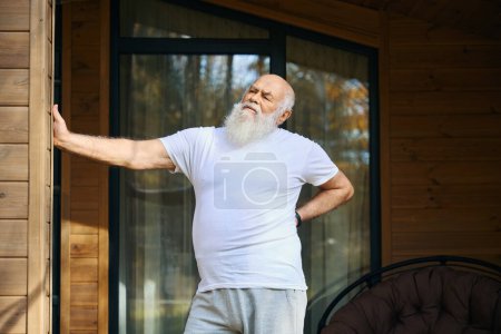Foto de Viejo sufre de dolor de espalda, se para en la veranda de una casa de campo - Imagen libre de derechos