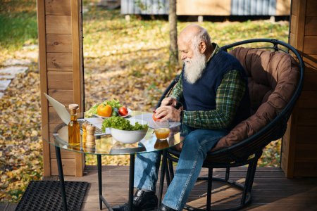 Pensionista se sienta en la terraza con el desayuno y un ordenador portátil, se sienta en una silla acogedora