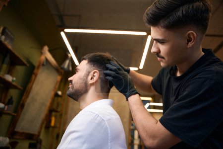 Téléchargez les photos : Barbier qualifié avec ciseaux brandissant artisanat coiffure élégante pour son client dans le salon de coiffure - en image libre de droit