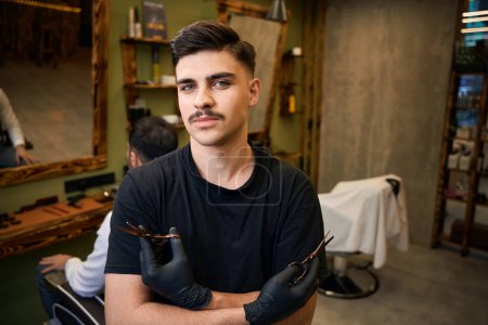 Téléchargez les photos : Barbier professionnel debout avec des bras croisés tenant des ciseaux dans le salon de coiffure - en image libre de droit