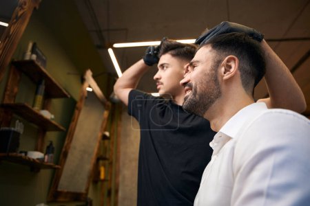 Téléchargez les photos : Barbier utilisant des ciseaux pour coiffer les cheveux de son client dans le salon de coiffure professionnel - en image libre de droit