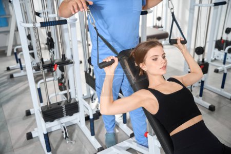 Téléchargez les photos : Femme faisant des exercices sur une machine sous la supervision d'un physiothérapeute, en utilisant un équipement moderne - en image libre de droit