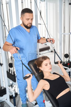 Téléchargez les photos : Femme fait des exercices pour renforcer ses muscles du dos sous la supervision d'un physiothérapeute à l'aide d'une machine d'exercice spéciale - en image libre de droit