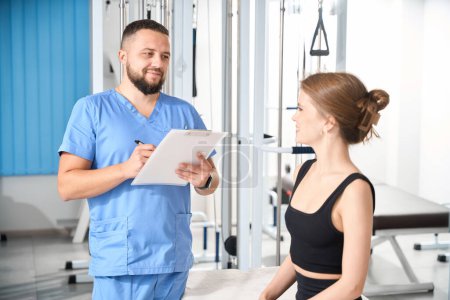 Photo for Kinesiologist in the clinic questions a young woman, the consultation takes place in the gym - Royalty Free Image