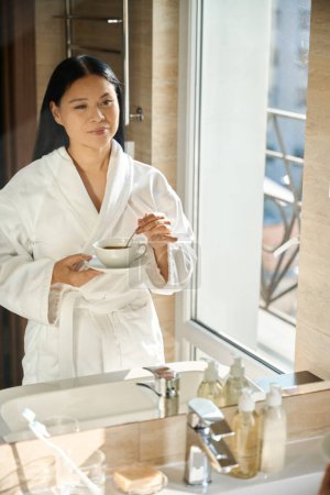 Ruhige Frau im Bademantel rührt koffeinhaltiges Getränk in Tasse mit Metallteelöffel, während sie vor dem Badezimmerspiegel steht