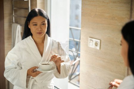 Porträt einer fokussierten Dame im Bademantel, die vor dem Spiegel Kaffee in Tasse mit Löffel rührt