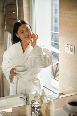 Photo for Smiling woman in bathrobe holding cup of coffee in hand while touching her face in front of mirror - Royalty Free Image