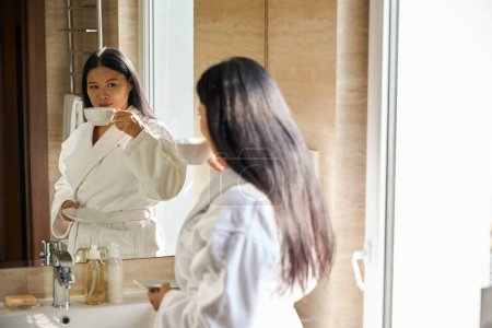 Téléchargez les photos : Femme agréable en peignoir tenant soucoupe à la main tout en soulevant la tasse à sa bouche devant le miroir - en image libre de droit