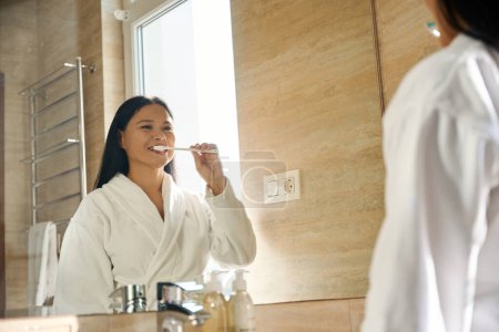 Téléchargez les photos : Femme en peignoir se brossant les dents avec une brosse à dents tout en se regardant dans un miroir - en image libre de droit