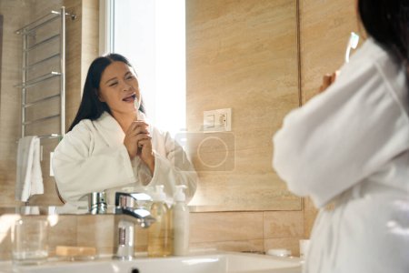 Téléchargez les photos : Dame vêtue de peignoir chantant tout en se brossant les dents avec une brosse à dents devant le miroir de salle de bain - en image libre de droit