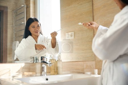 Téléchargez les photos : Joyeuse femelle vêtue de peignoir pointant sa brosse à dents vers son reflet dans le miroir de salle de bain - en image libre de droit