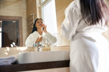 Téléchargez les photos : Femme en peignoir appliquant de la crème cosmétique sur son visage tout en regardant son reflet dans le miroir - en image libre de droit