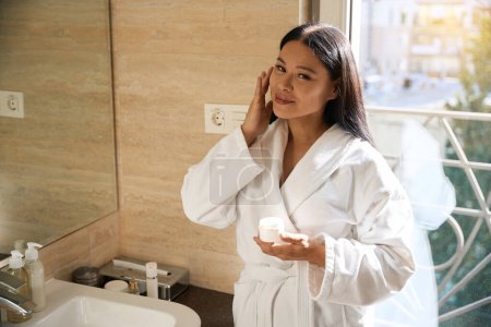 Téléchargez les photos : Femme satisfaite en peignoir appliquant de la crème cosmétique sur son visage tout en se tenant devant le miroir de salle de bain - en image libre de droit