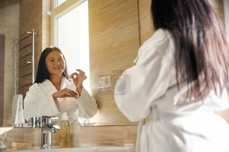 Foto de Mujer alegre en albornoz goteando aceite cosmético en la parte posterior de su mano delante del espejo del baño - Imagen libre de derechos