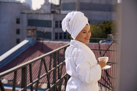 Téléchargez les photos : Dame agréable en peignoir avec tasse de boisson caféinée et soucoupe dans les mains posant pour caméra sur le balcon donnant sur le paysage urbain - en image libre de droit