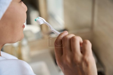 Photo for Closeup of lady in white terry bathrobe and bath towel wrapped around head lifting toothbrush to her mouth - Royalty Free Image