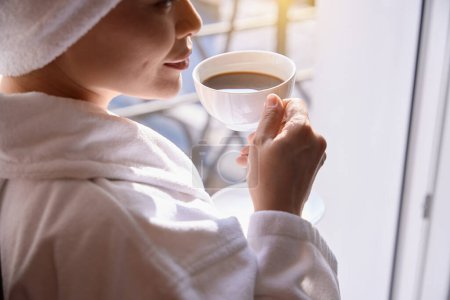 Foto de Primer plano de la señora en bata de baño de rizo blanco sosteniendo platillo en la mano mientras levanta la taza de espresso a la boca - Imagen libre de derechos