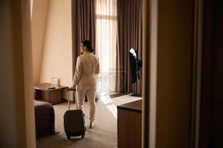 Téléchargez les photos : Vue arrière de la femme adulte avec valise trolley debout au milieu de sa suite - en image libre de droit