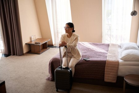 Photo for Smiling happy female tourist seated on bed in her suite leaning on trolley suitcase handle - Royalty Free Image