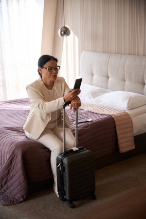 Téléchargez les photos : Femme d'affaires assise sur le lit dans la chambre d'hôtel appuyée sur la poignée de la valise tout en regardant l'écran du smartphone - en image libre de droit