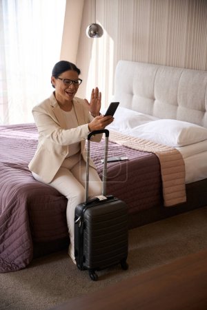 Téléchargez les photos : Gai dame d'affaires assis sur le lit en suite avec valise agitant quelqu'un sur son écran de smartphone - en image libre de droit