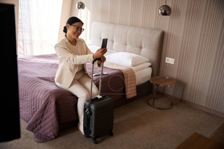 Foto de Mujer sonriente sentada en la cama en la habitación de hotel apoyada en el asa de la maleta de viaje mirando el teléfono celular en su mano - Imagen libre de derechos