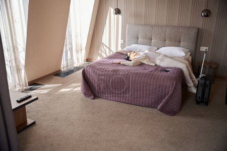 Téléchargez les photos : Voyageuse fatiguée avec smartphone à la main et documents de voyage couchés sur le lit dans sa suite de luxe - en image libre de droit
