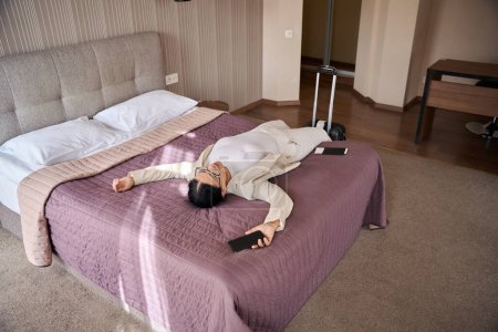 Foto de Mujer de negocios cansada con teléfono móvil en la mano y documentos de viaje durmiendo en la cama en su suite - Imagen libre de derechos