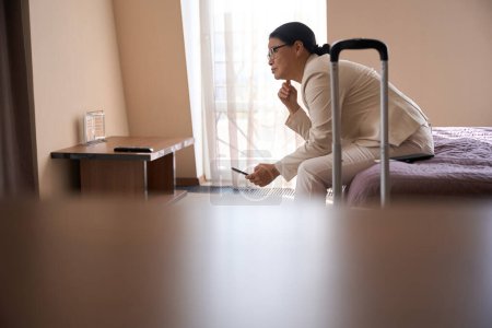 Téléchargez les photos : Femme d'affaires réfléchie avec smartphone à la main assise sur le lit dans la chambre d'hôtel et regardant au loin - en image libre de droit
