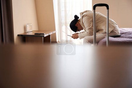 Exhausted business lady with cellular phone in hands sitting on edge of bed in hotel room