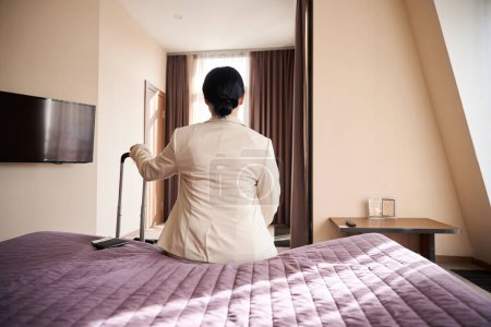 Téléchargez les photos : Vue arrière de la femme assise sur le bord du lit dans la chambre d'hôtel tout en s'appuyant sur la poignée de la valise - en image libre de droit