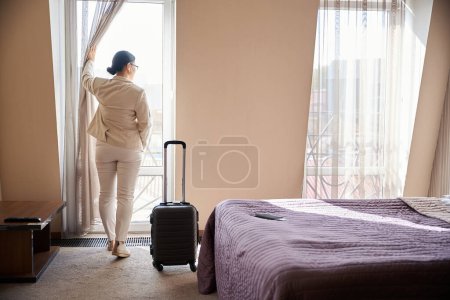 Photo for Back view of woman looking through curtained balcony door while standing in her hotel room - Royalty Free Image