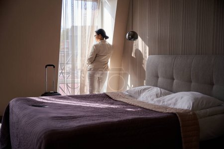 Téléchargez les photos : Vue arrière de la femme regardant par la fenêtre ridée dans sa chambre d'hôtel - en image libre de droit