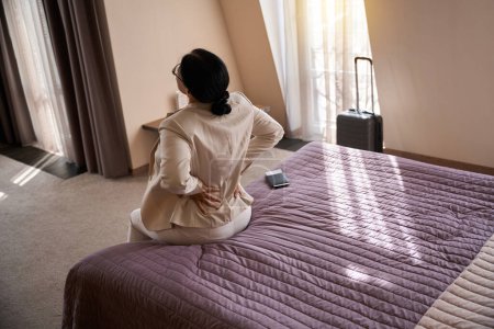 Téléchargez les photos : Femme assise sur le bord du lit en suite avec les mains sur le bas du dos - en image libre de droit