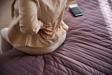 Téléchargez les photos : Photo recadrée de dame touchant son bas du dos avec les deux mains tout en étant assis sur le lit dans la chambre d'hôtel - en image libre de droit