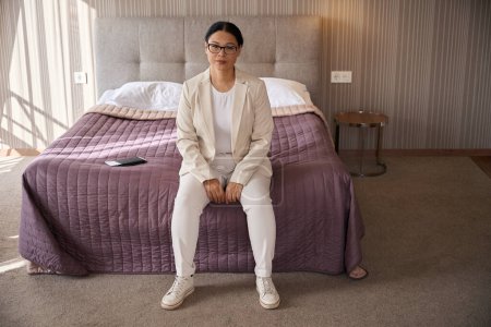 Photo for Calm elegant mature woman in eyeglasses sitting on edge of bed in hotel room - Royalty Free Image