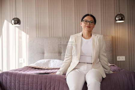 Téléchargez les photos : Dame satisfaite dans les lunettes assis sur le bord du lit dans sa suite - en image libre de droit