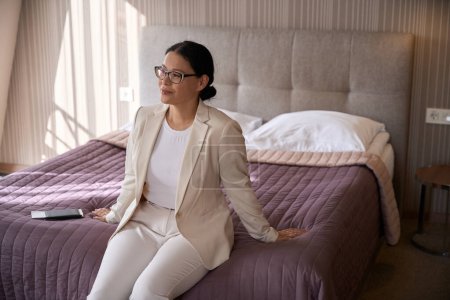 Téléchargez les photos : Agréable femme d'affaires en lunettes assise sur le bord du lit dans la chambre d'hôtel regardant loin - en image libre de droit