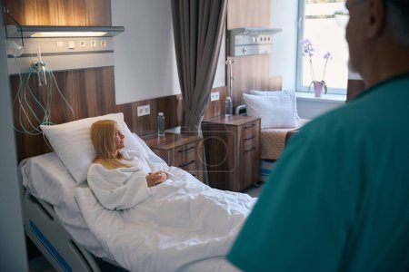 Attending physician standing in front of female patient lying in bed in hospital room