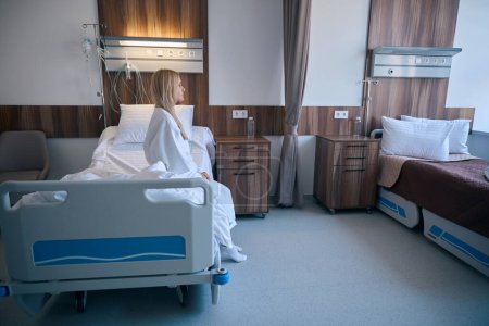 Foto de Vista lateral del triste paciente adulto hospitalizado en albornoz sentado en el borde de la cama en la sala de instalaciones médicas - Imagen libre de derechos