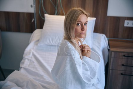 Photo for Waist-up portrait of dejected scared female patient dressed in bathrobe seated on bed in hospital ward - Royalty Free Image