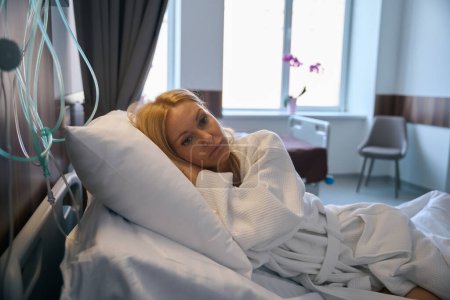 Traurige nachdenkliche Frau im Bademantel liegt in Krankenhausstation im Bett